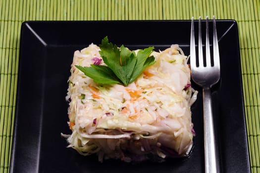 Mixed cabbage salad with fork is served on black square shape plate on green background.