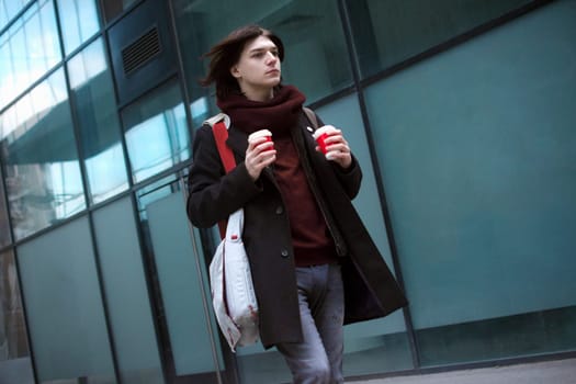 Trendy young guy with laptop bag and two coffees is walking or hurrying at the street. Work day morning.