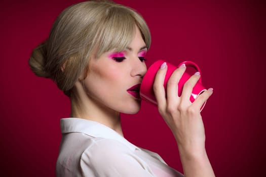The fashionable trendy blond hair woman with pink make-up is drinking from red or pink mug on red background.