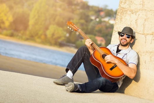 The cheerful singing hipster guy is playing guitar and sitting outdoors at the riverside.