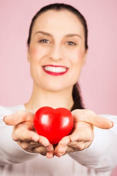 The smiling woman is holding and giving red heart and sharing love on pink background.