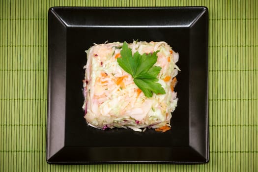 Mixed cabbage salad with parsley decoration is on black plate on green tablecloth.