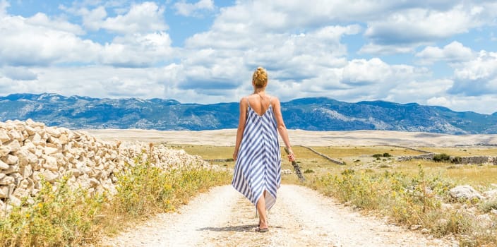 Rear view of woman in summer dress holding bouquet of lavender flowers while walking outdoor through dry rocky Mediterranean coast lanscape on Pag island, Croatia in summertime.