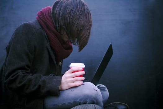 The hacker or freelancer person with coffee in hand is using laptop on dark background.