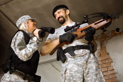 Smiling cheerful male and female military persons are holding crossbow and binoculars indoors.
