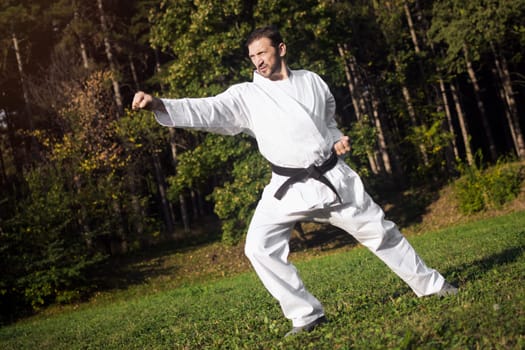 The middle aged man in white kimono and black belt is practicing martial arts in nature.