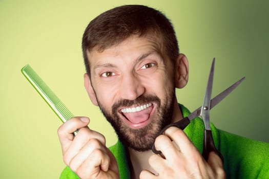 Smiling cheerful male hairdresser is holding a comb and scissors in hands on yellow green background, haircut for men concept.