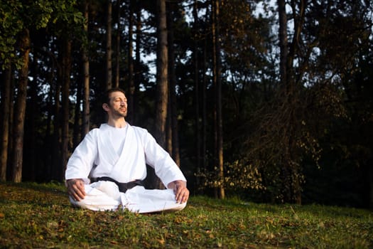 Man in white kimono with black belt is practicing meditation in forest in nature.