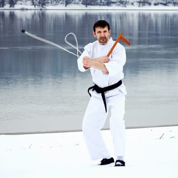 The man in white kimono and black belt is practicing using kusarigama weapon on snow at winter.