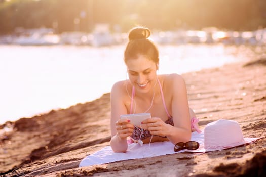 The smiling woman is using phone and sunbathing on the beach.