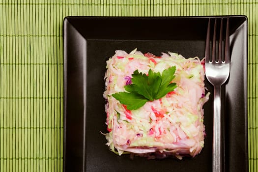 The mixed cabbage salad with fork is served on the black square plate on green background.
