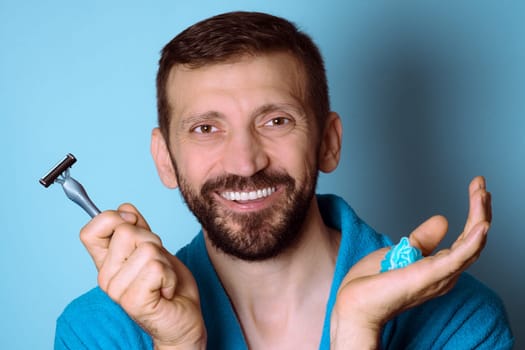 The smiling bearded man is ready for shaving and holding a razor and shaving gel in hand on blue background.