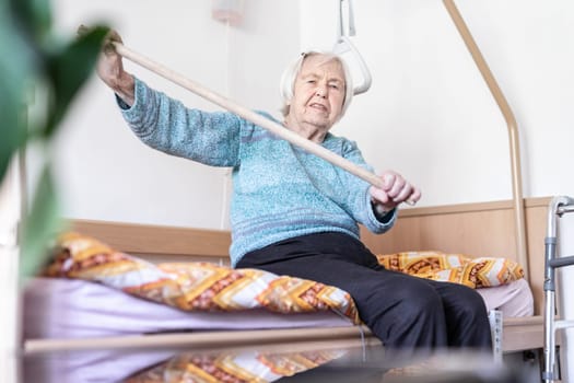 Elderly 96 years old woman exercising with a stick sitting on her bad. Geriatric health care home assisted support for older people concept. Care for the elderly.