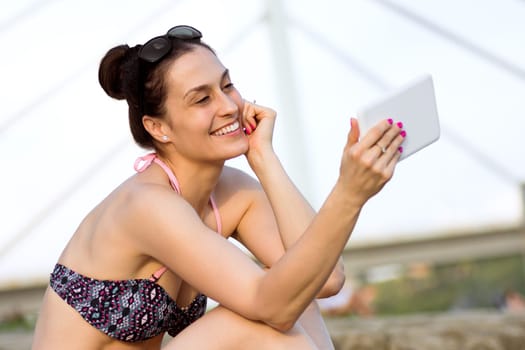 The smiling woman in bikini is using digital tablet on the beach.