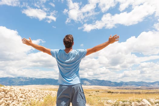 Rear view of casual sporty man standing on a dirt country road rising hands up to the clouds on a blue summer sky. Freedom and travel adventure concept. Copy space.