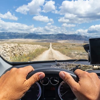 Detail of male driver hands on steering wheel. Driving a car on country road on dramatic countryside lanscape of Pag island, Croatia. View from the cabin trough the windshield.