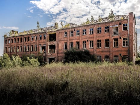 Industrial building covered with shrubs. Ruin with sunken roof against cloudy blue sky