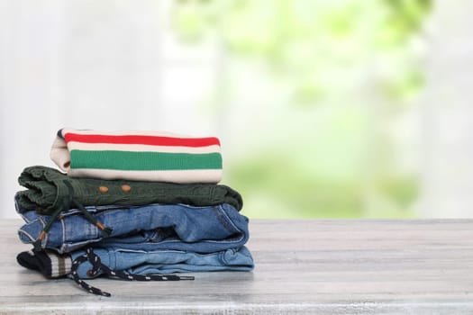 Stack of jeans. Closeup of a pile of colorful denim pants and one colorful sweater on a bright table against abstract blurred natural light green background. Space for your product display montage.