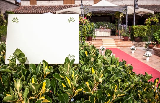 Wedding invitation sheet placed over green hedge with wedding reception as background