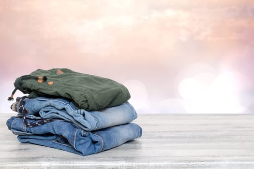 Stack of jeans. Closeup of a pile of colorful denim pants on a bright table against abstract blurred natural light pink background. Space for your product display montage.