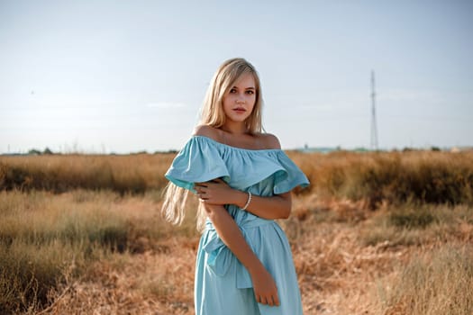 young beautiful caucasian blonde girl in light blue dress stands on the field with the sun-scorched grass