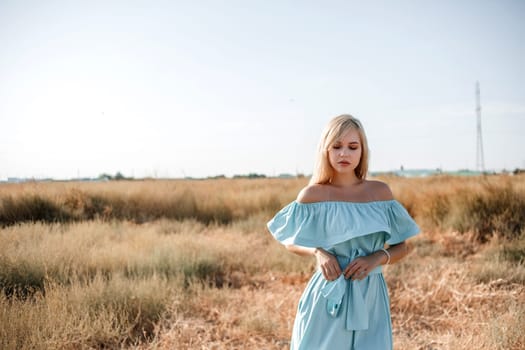 young beautiful caucasian blonde girl in light blue dress stands on the field with the sun-scorched grass