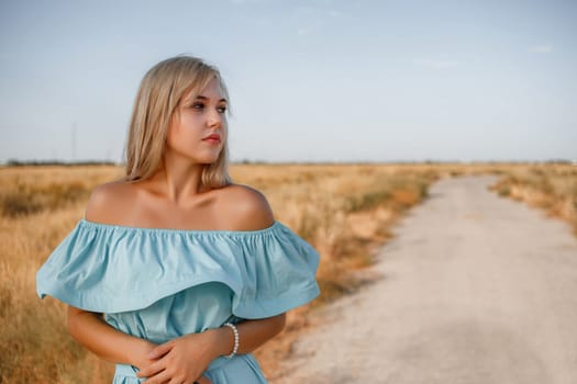 portrait of a young beautiful caucasian blonde girl in a light blue dress standing on a field with sun-dried grass next to a small country road