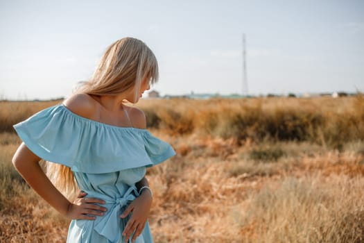 young beautiful caucasian blonde girl in light blue dress stands on the field with the sun-scorched grass