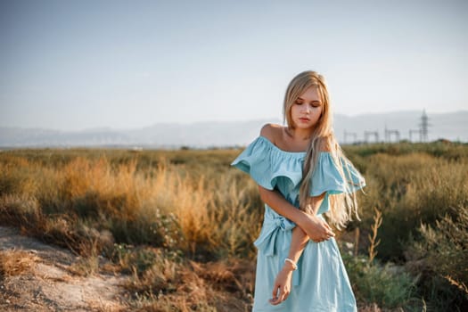 portrait of a young beautiful caucasian blonde girl in a light blue dress standing on a field with sun-dried grass next to a small country road