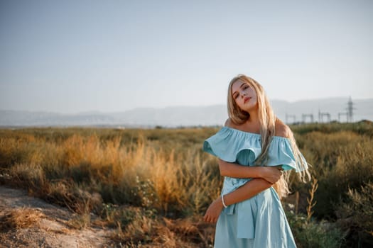 portrait of a young beautiful caucasian blonde girl in a light blue dress standing on a field with sun-dried grass next to a small country road