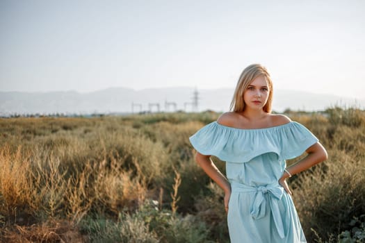 portrait of young beautiful caucasian blonde girl in light blue dress standing on the field with sun-dried grass during sunset