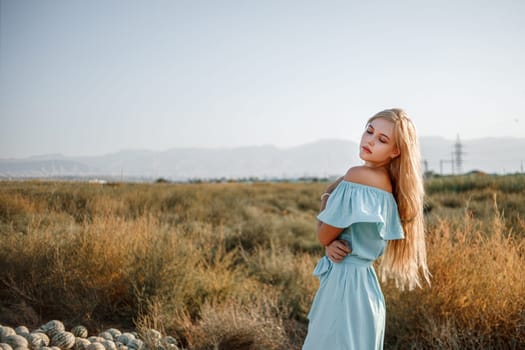 portrait of a young beautiful caucasian blonde girl in a light blue dress standing on a watermelon field with sun-dried grass during sunset