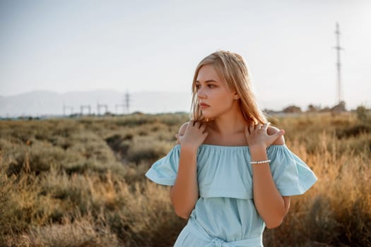 portrait of young beautiful caucasian blonde girl in light blue dress standing on the field with sun-dried grass during sunset