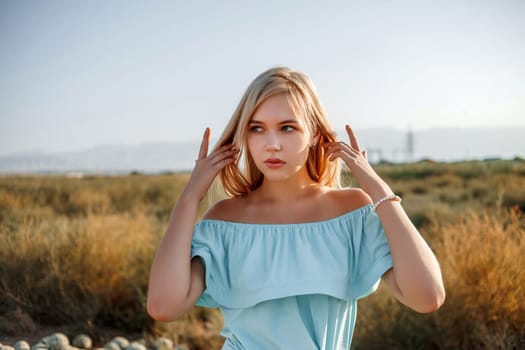 portrait of a young beautiful caucasian blonde girl in a light blue dress standing on a watermelon field with sun-dried grass during sunset