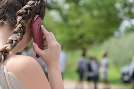 Close-up over the shoulder young girl talking on the mobile phone with green foliage and blurry people on background.