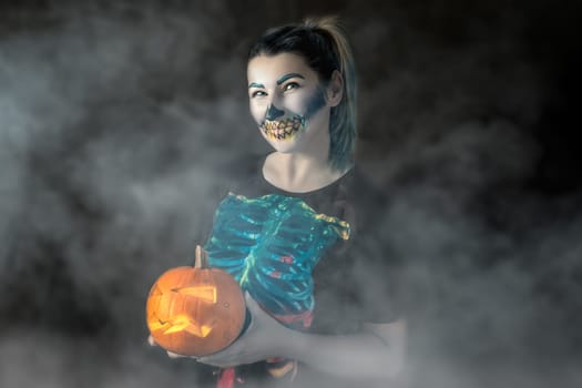 Halloween portrait, adult Caucasian white woman holding orange pumpkin, dressed and makeup stylised for skeleton on dark foggy or misty background