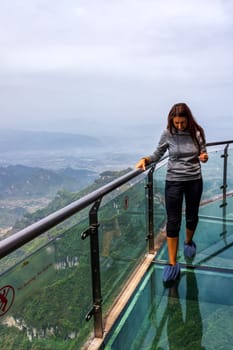 Young Caucasian white woman walking on glass walkway, Sky Walk at Tianmen Mountain, view on valley and mountains down below, Zhangjiajie, Hunan, China