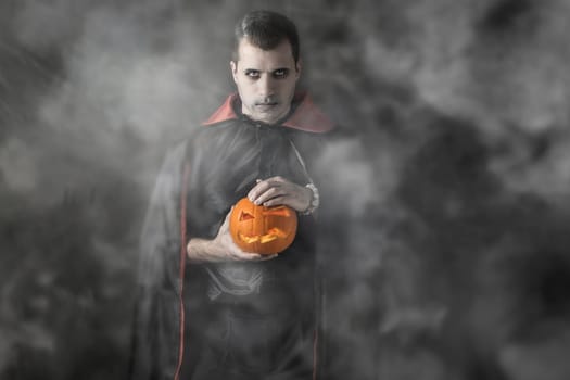 Halloween portrait, adult Caucasian white man holding orange pumpkin dressed and makeup stylised for a vampire on dark smoky foggy or misty background