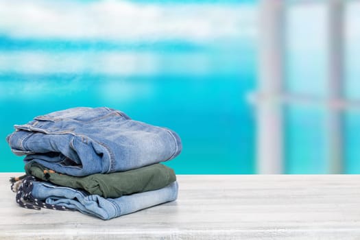 Stack of jeans. Closeup of a pile of colorful denim pants on a bright table against abstract blurred light blue windows background. Template for your product display montage.