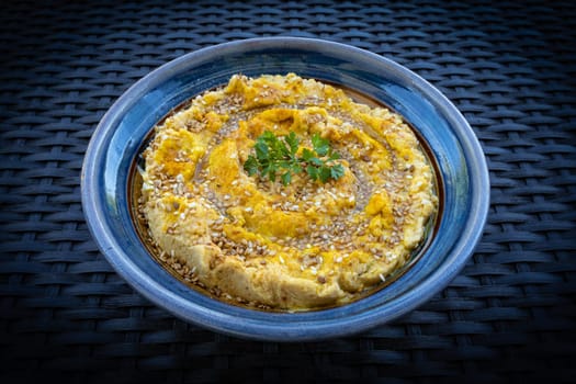 Tasty hummus (or houmous) in blue bowl on dark background