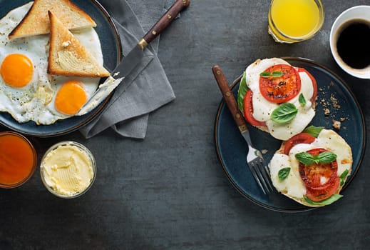 Breakfast served with coffee, jam, butter, mozzarella sandwich and fried eggs with bread on dark table top view. Heathy breakfast food.