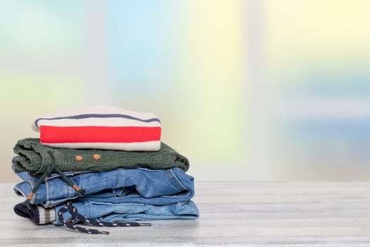 Stack of jeans. Closeup of a pile of colorful denim pants and one colorful sweater on a bright table against abstract blurred natural light pastel background. Space for your product display montage.