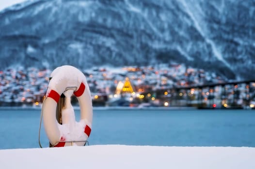 Lifebuoy covered in snow on the shore of famous Tromso Bridge across Tromsoysundet strait, Northern Norway