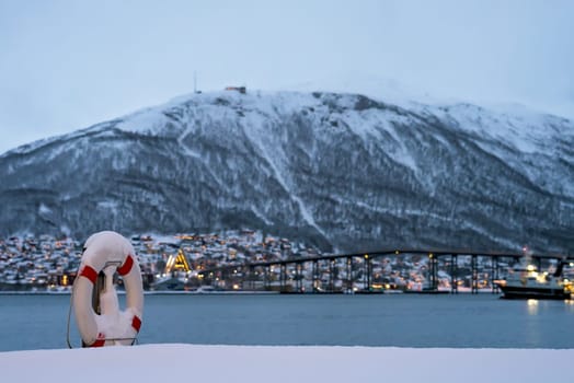 Lifebuoy covered in snow on the shore of famous Tromso Bridge across Tromsoysundet strait, Northern Norway