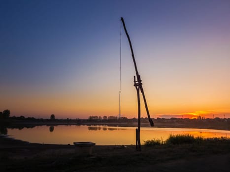 Idyllic rural scene with an old water well (popular called the stork well in the old villages of Moldova, also well sweep or shadoof). Calm sunset sky reflecting on the lake water surface.