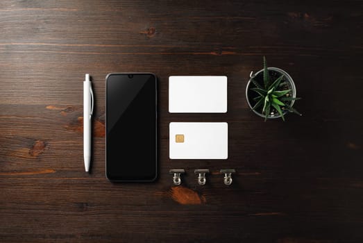 Blank business template. Smartphone, business card, bank card, pen and plant on wood table background. Flat lay.