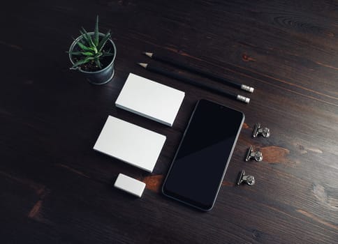 Photo of blank stationery and cellphone on wood table background. Smartphone, business cards, pencils, eraser and plant.