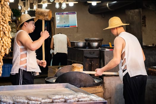 Xian, China -  July 2019 : Two men hitting hard with big wooden hammers to crack grain which will be used in making of sweet snack for sale on the street, Muslim Quarter