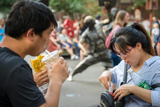 Xian, China -  July 2019 : Man drinking fruit juice from a plastic cup through a straw and woman looking for something in her handbag on the street in the Muslim Quarter