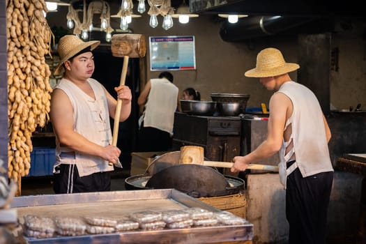 Xian, China -  July 2019 : Two men hitting hard with big wooden hammers to crack grain which will be used in making of sweet snack for sale on the street, Muslim Quarter